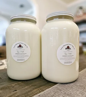 Two large glass jars of raw milk from Tanglewood Farm, displayed on a wooden table. Each jar features a label with the farm's logo and product information, highlighting the freshness and quality of the milk.